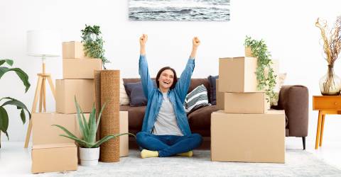 Vrolijke jonge vrouw zit blij op de grond tussen haar verhuisdozen in haar nieuwe huis